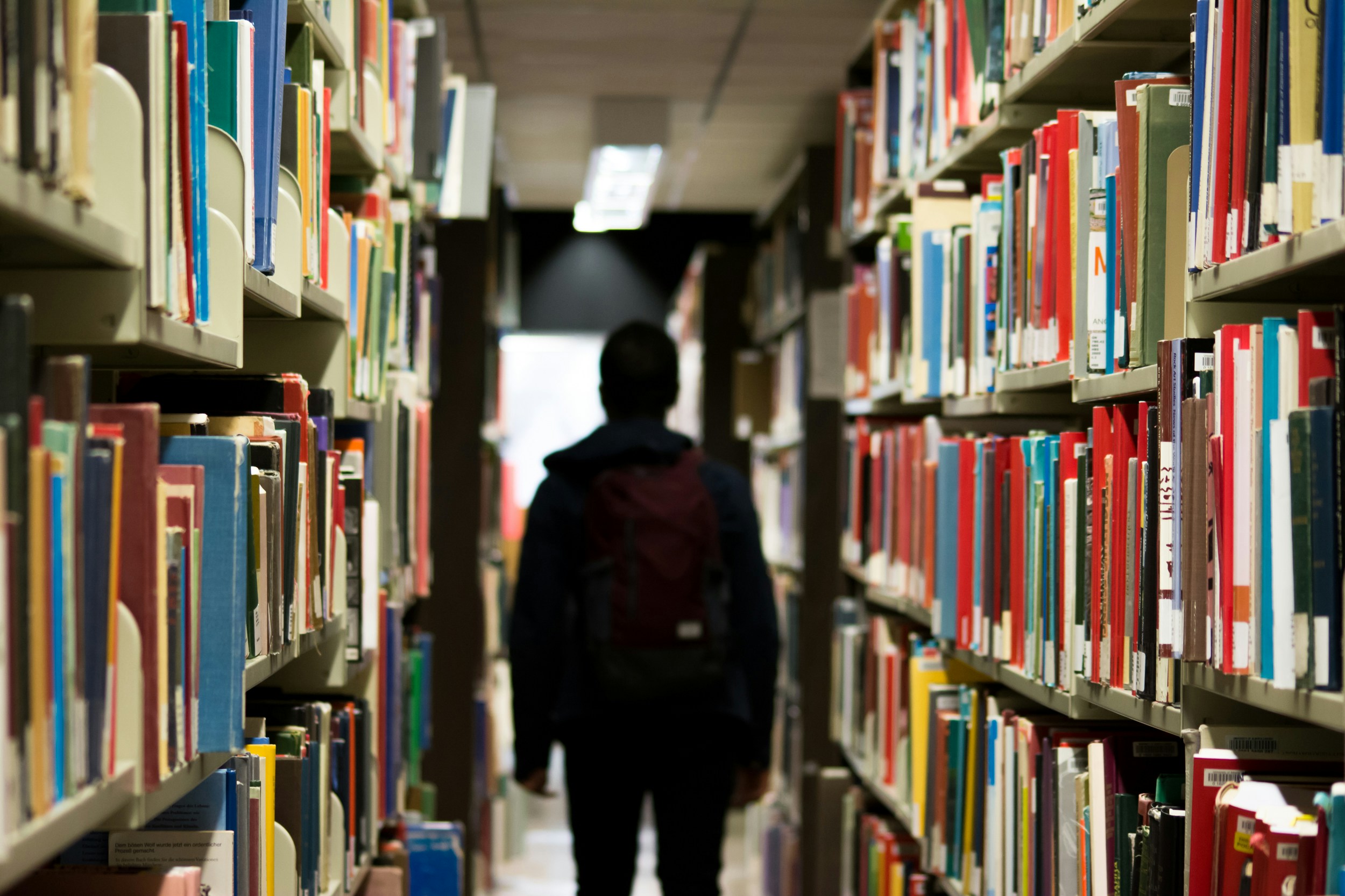 Shadow of a man walking through rows of library bookshelves, creating a silhouette against the dimly lit aisle filled with books.