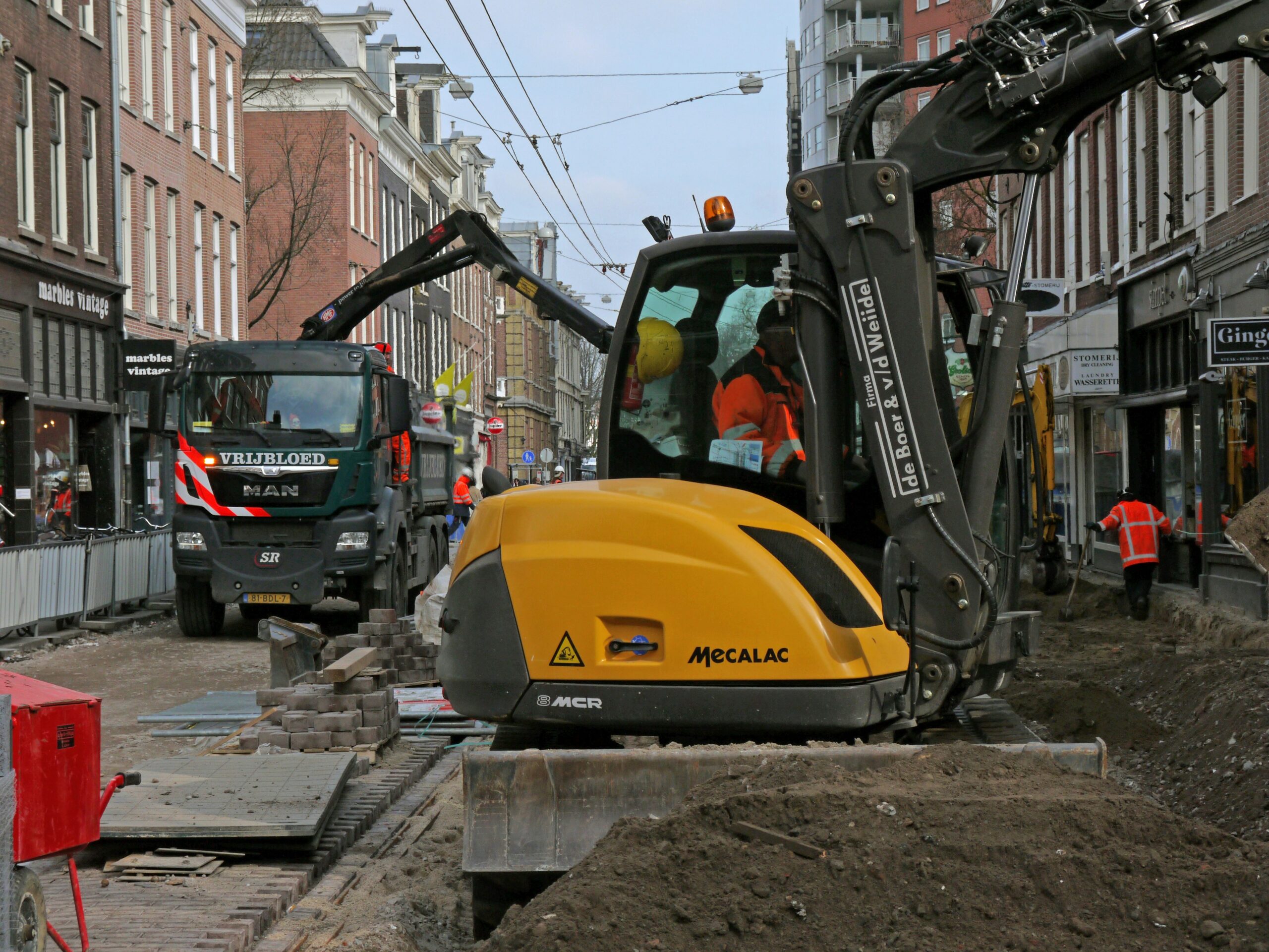 Construction site in a market town with diggers and cranes actively working on new developments. The scene includes partially built structures surrounded by temporary fencing and construction materials, set against a backdrop of existing town buildings.